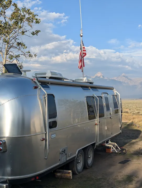A silver camping trailer with an antenna mast and an American flag against distant mountains.