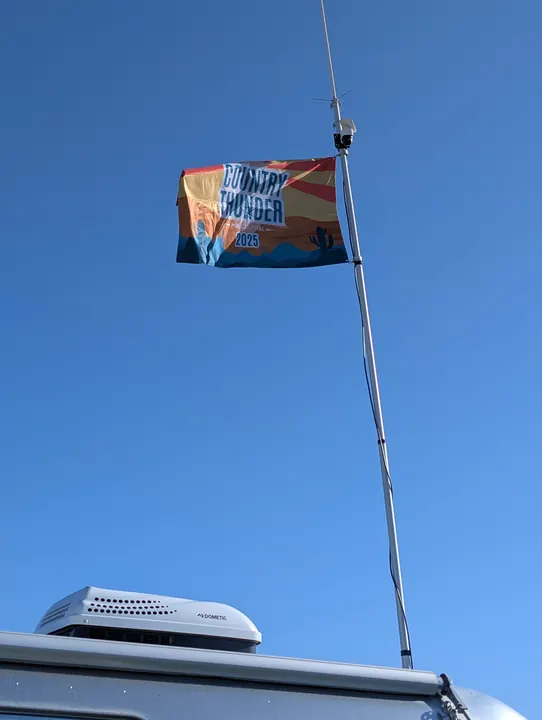 An antenna mast and colorful festival flag above a camper roof against a clear blue sky.