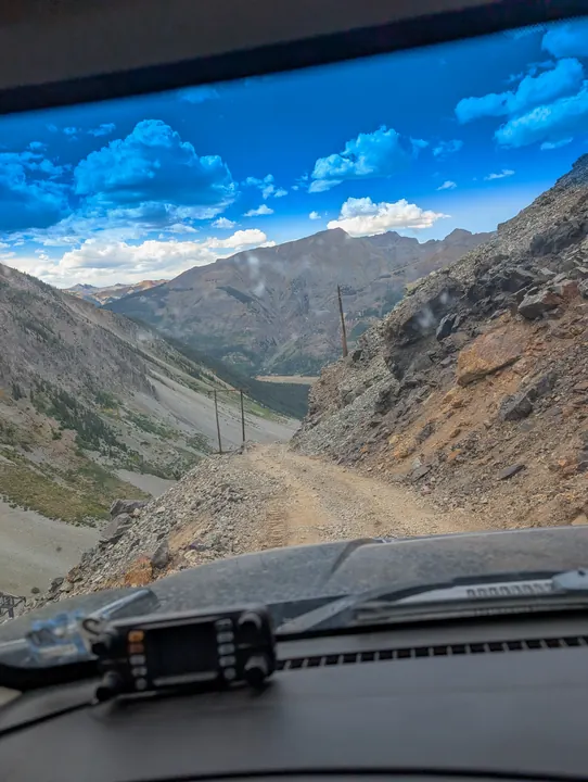 A dashboard radio overlooking a rough mountain road descending toward a lake between steep ridges.