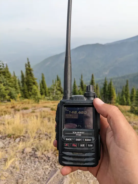 A handheld radio above a grassy mountain ridge with evergreen trees and hazy peaks.