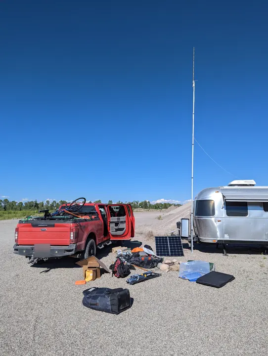 A tall portable antenna mast, solar panel, and camping equipment beside a red truck and silver trailer.