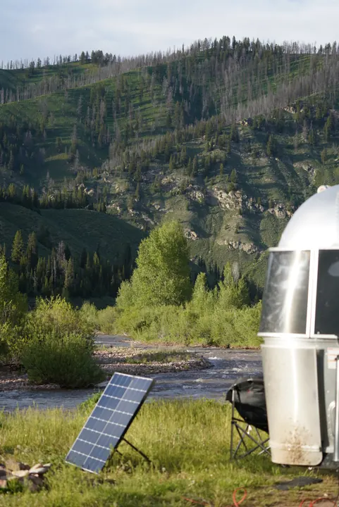 A portable solar panel and camp chair beside a silver trailer, with a river and wooded hillside behind.