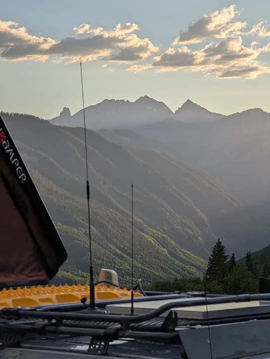 Vehicle radio antennas in front of layered mountain ridges lit by the setting sun.