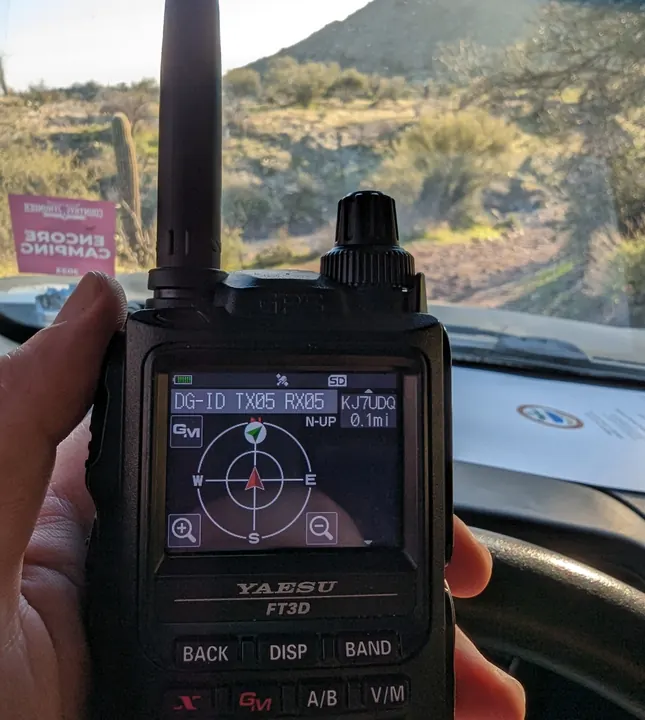 A handheld radio displaying its direction screen inside a vehicle, with a desert hillside outside.